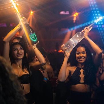 Women in a nightclub holding bottles with sparklers, celebrating bottle service under neon lights.