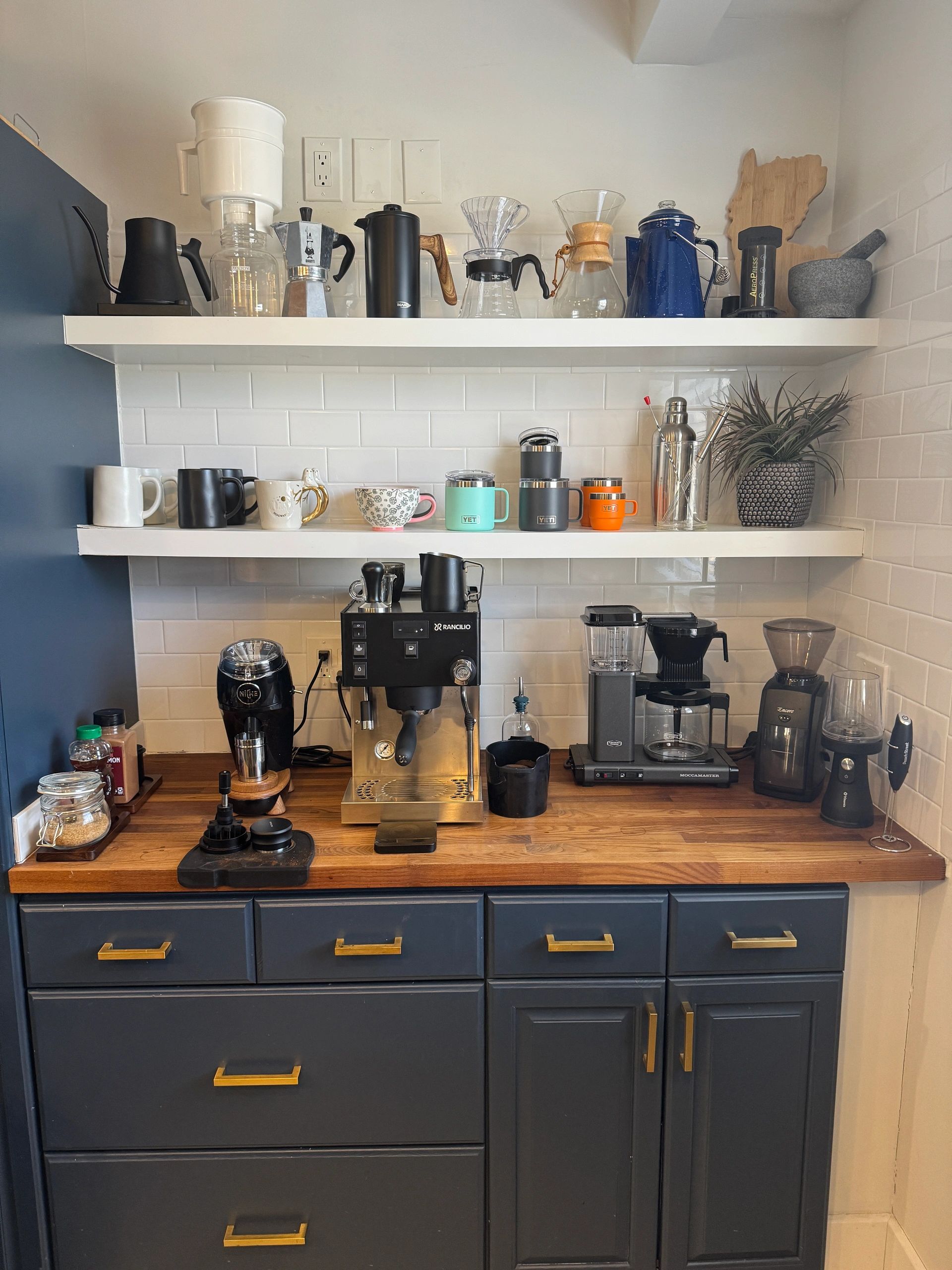 Organized coffee station with espresso machine, grinders, mugs, and brewing tools on wooden counter and white shelves.