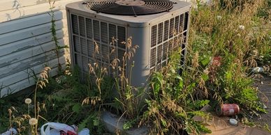 Overgrown weeds and trash surround an outdoor air conditioning unit by a house.