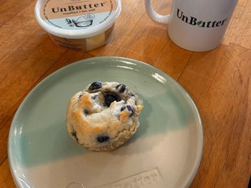 Blueberry scone on a plate with butter and a mug on a wooden table.