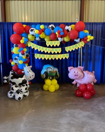 Red, blue, yellow, and cow print balloon arch. Large cow, tractor and pig balloons in front.