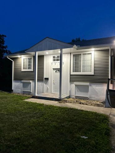 Well-lit modern house entrance at night with gray siding and white trim.