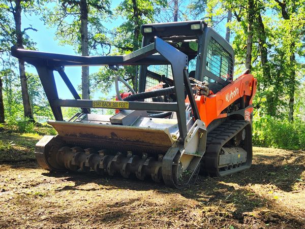 Kubota skid-steer loader with forestry mulcher attachment in a wooded area.