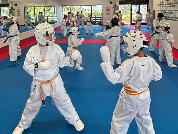 Children sparring in a karate class wearing protective gear and orange belts.