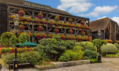 The Dickens Inn adorned with vibrant flowers on its balconies and surrounded by lush greenery.
