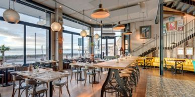 Modern restaurant interior with wooden tables and large windows overlooking the sea.