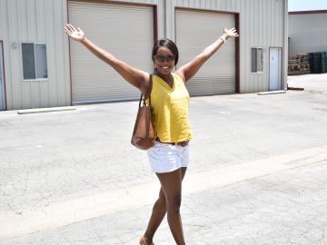 Woman in yellow top and white shorts joyfully posing outdoors with arms raised.