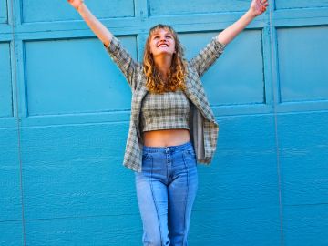A joyful woman with arms raised stands against a bright blue wall.