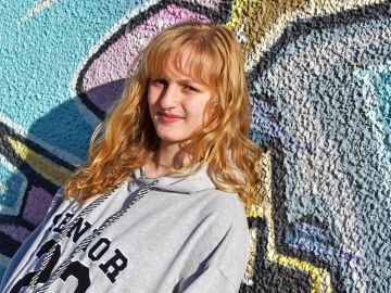 Young woman with red hair in a gray hoodie stands against colorful graffiti wall.