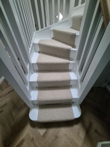 Narrow carpeted staircase with white railings and beige runner secured by stair rods.
