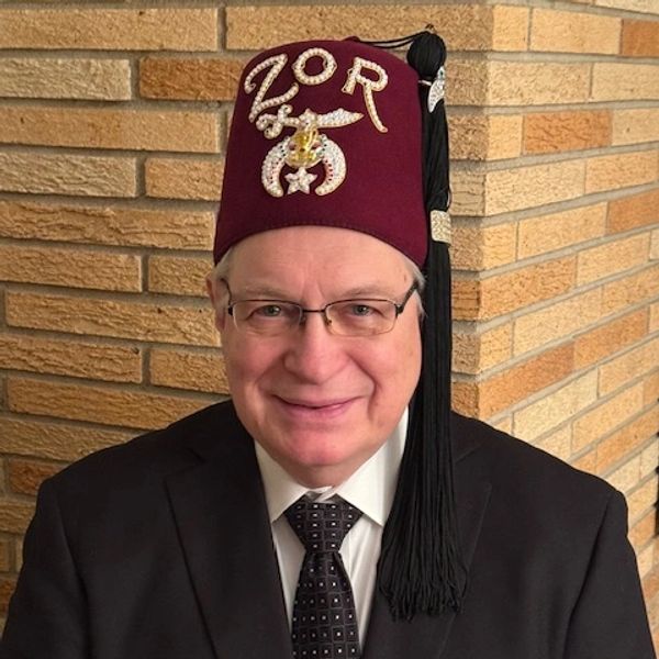 Man in glasses and suit wearing a maroon fez with decorative lettering.