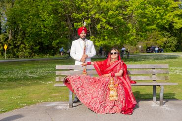 A bride in a red traditional dress sitting on a bench, with a groom standing behind her in a white suit and red turban.