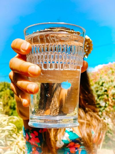 Hand holding a glass of water outdoors with blue sky background.