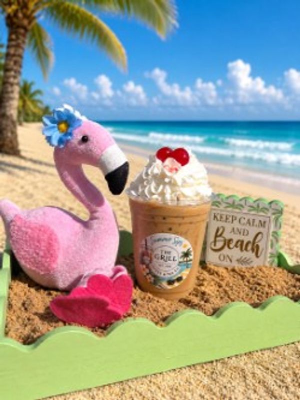 A plush pink flamingo, iced coffee, and a beach sign on a sandy tray by the ocean.