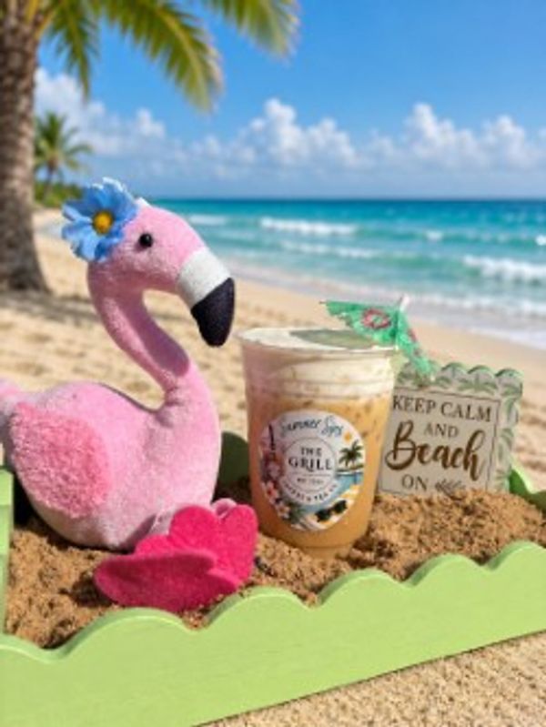 A pink flamingo toy, iced coffee, and a beach sign on sandy shore with ocean background.