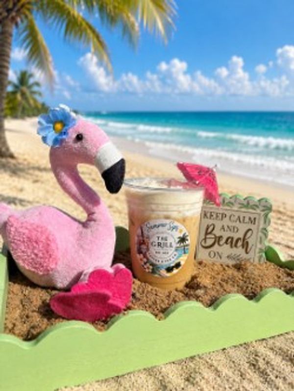 Pink flamingo plush with iced drink and beach sign on sandy shore.