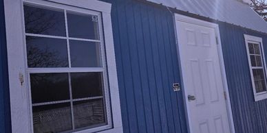 Blue building with white door and windows under a corrugated metal roof.