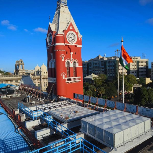 Red clock tower with Indian flag in urban setting under clear blue sky.