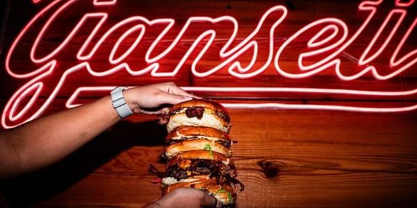 Person holding a towering stacked burger in front of a red neon sign.