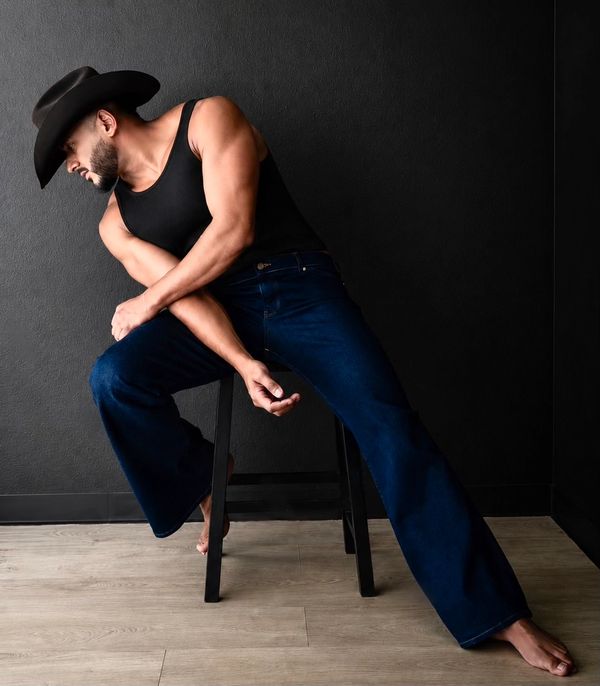Man in cowboy hat and tank top sitting on stool.