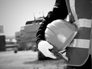 Black and white image of a person holding a hard hat while wearing white gloves and a safety vest