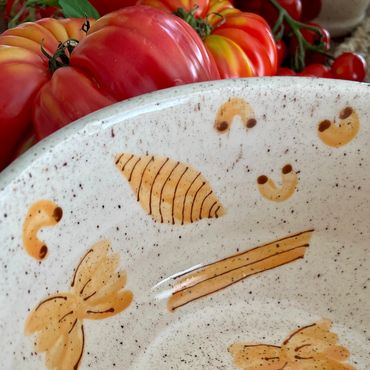 A close up detail show of the hand painted pastas on a speckeled ceramic bowl with tomatos.