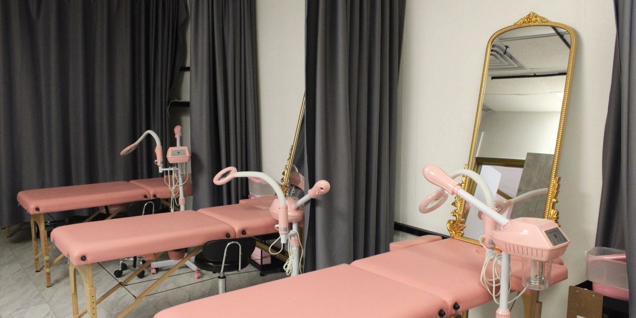 Pink treatment beds and machines in a beauty clinic room with mirrors and dark curtains.