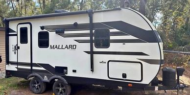 A white and black Mallard travel trailer parked outdoors among trees.