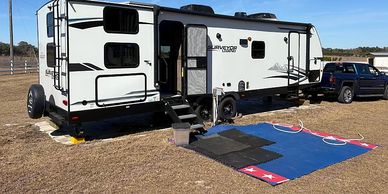 A white recreational vehicle parked on a grassy area with a blue mat outside.