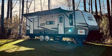 A Coleman travel trailer parked in a wooded area with sunlight filtering through trees.