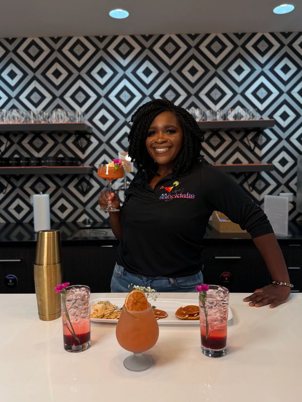 Smiling woman holding a cocktail behind a bar with colorful drinks and appetizers.