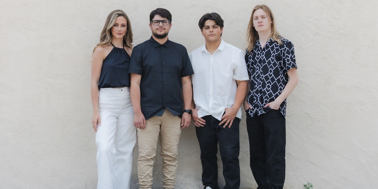 Four young adults standing against a beige wall, casually dressed.