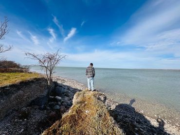 A person stands on a rocky edge overlooking a calm body of water under a blue sky.