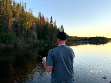 Man fishing alone by a calm lake surrounded by trees at sunset.