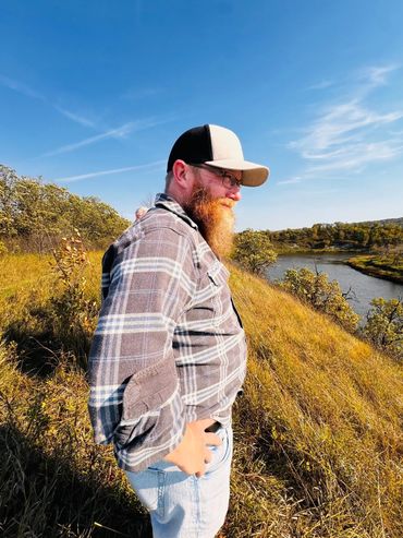 Man with a beard and cap standing on a grassy hill by a river.