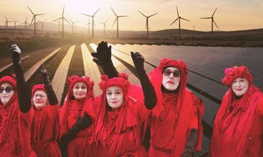Seven people in red robes pose in front of solar panels and wind turbines at sunset.
