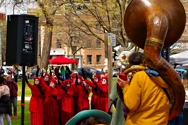 Performers in red costumes with white face paint raising hands during an outdoor event.