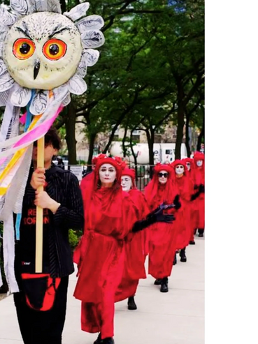 People in red robes march behind a person holding a large owl-faced flower puppet.