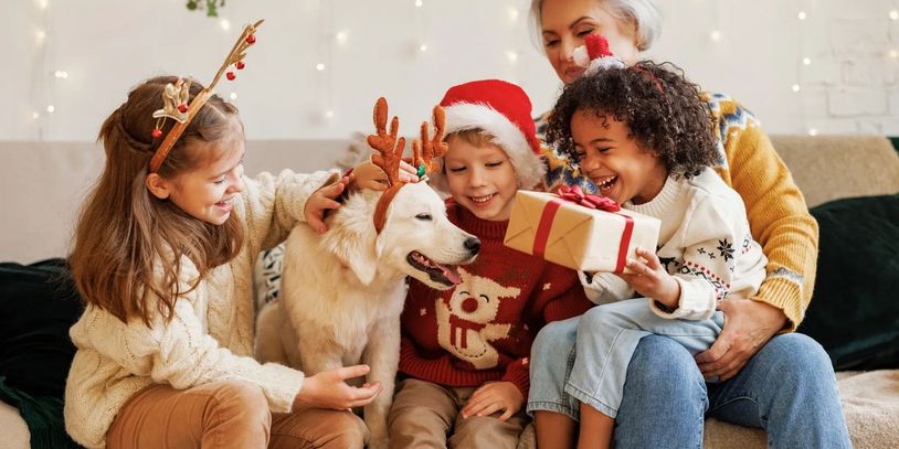 Children and an elderly woman celebrate Christmas with a dog wearing reindeer antlers and a gift.