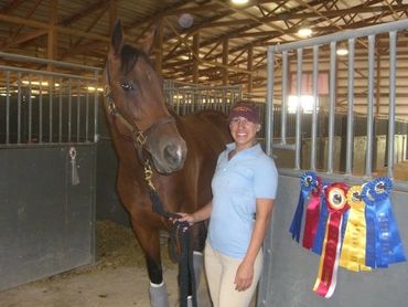 Woman standing in front of a horse stall with winning ribbons, holding a brown horse. 