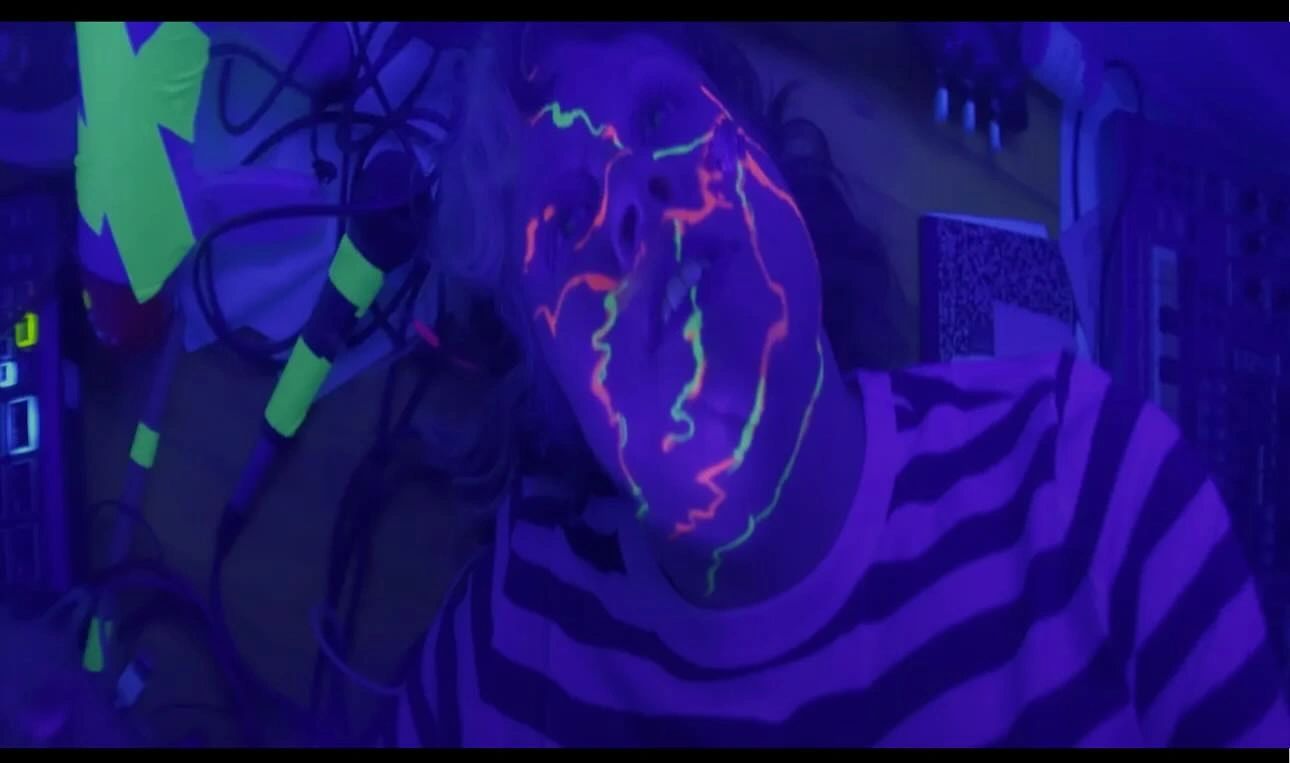Person with neon paint on face under blacklight in a striped shirt.