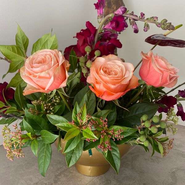 Modern floral arrangement in gold vase with apricot roses, green leaves.