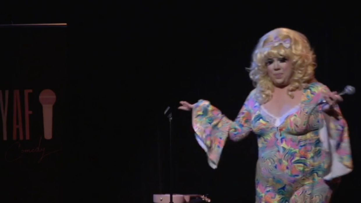 Comedian in a colorful dress performing on stage with a microphone.