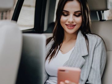 Woman smiling while using phone and laptop in a car.