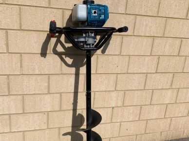 Gas-powered earth auger leaning against a brick wall.