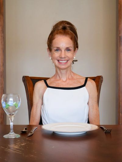 Smiling woman in a white dress sitting at a dining table with an empty plate and glass.