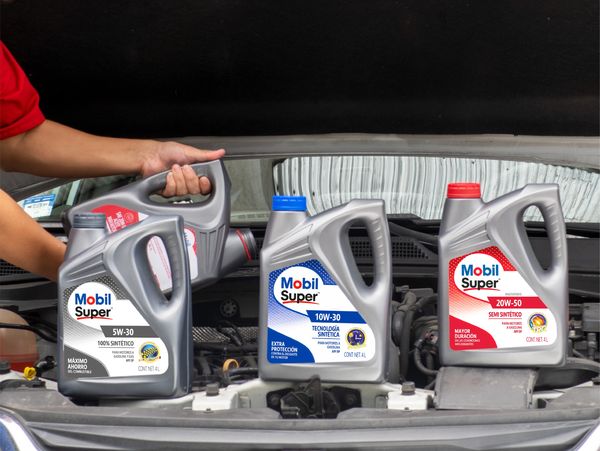 Three Mobil Super motor oil containers placed on a car engine with a person holding one.