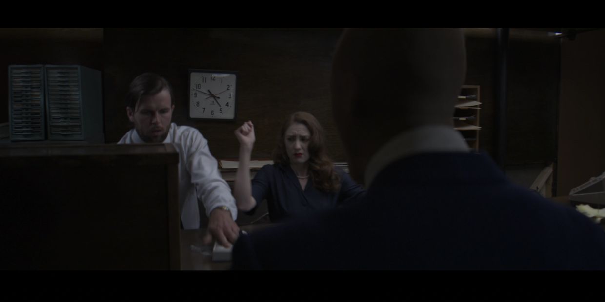 Two people sit at a desk, apprehensive, while another watches in a dim office.
