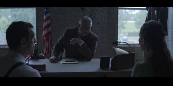 Three people in a serious discussion in a dimly lit office with an American flag.