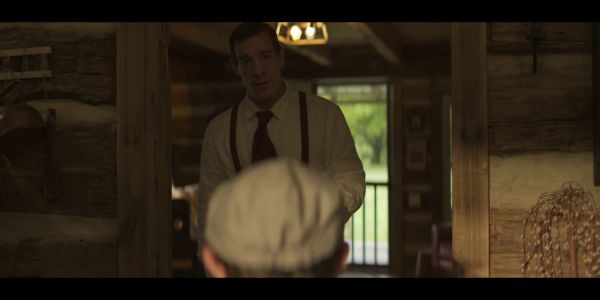 Man in vintage attire faces another inside a rustic wooden cabin.